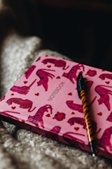 A notebook with a pink cover adorned with red tiger illustrations and small red hearts. The word 'notebook' is printed in the center. A pen with a tiger-striped design is placed on top of the notebook.