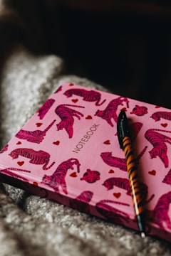 A notebook with a pink cover adorned with red tiger illustrations and small red hearts. The word 'notebook' is printed in the center. A pen with a tiger-striped design is placed on top of the notebook.