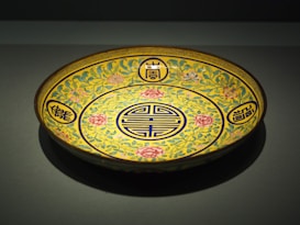 A decorative ceramic plate with intricate patterns, showcasing floral motifs in pink, green, and blue against a yellow background. Chinese characters are encircled in blue and placed symmetrically around the central design.