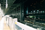 A sleek, modern high-speed train is stationed at a brightly lit platform. The station features an array of lights and structural beams overhead, surrounded by transparent glass walls. There is minimal movement, suggesting a calm and organized atmosphere.