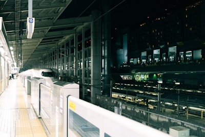 A sleek, modern high-speed train is stationed at a brightly lit platform. The station features an array of lights and structural beams overhead, surrounded by transparent glass walls. There is minimal movement, suggesting a calm and organized atmosphere.