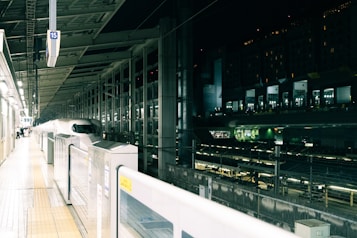 A sleek, modern high-speed train is stationed at a brightly lit platform. The station features an array of lights and structural beams overhead, surrounded by transparent glass walls. There is minimal movement, suggesting a calm and organized atmosphere.