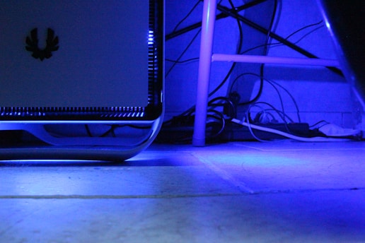 A computer tower with LED lighting is placed next to a desk. The room is illuminated with a blue hue, and various cables are visible behind the desk, creating a technological and somewhat cluttered appearance.