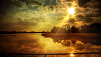 The image captures a serene lake scene at sunset with the sun casting a golden hue across the sky and water. Rays of sunlight break through scattered clouds, and the reflective surface of the lake mirrors the vibrant colors and textured sky. Trees and reeds line the shore, silhouetted against the brilliant backdrop.