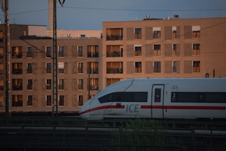 A high-speed train travels along tracks in front of modern apartment buildings. The sun is setting, casting a warm light on the buildings' facades. The train is white with a red stripe and is marked with a bicycle symbol, indicating bike-friendly facilities.