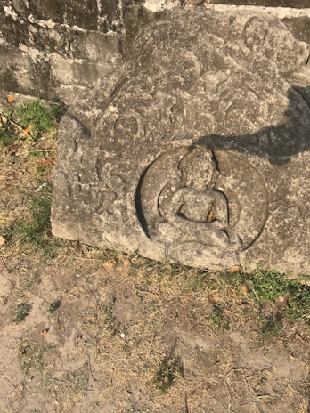 An ancient stone carving depicting a seated figure in a meditative pose, surrounded by trees and grass. The stone surface shows signs of weathering and age, with moss and greenery around it.