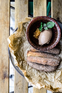 A top-down view of a wooden table with a plate containing churros dusted with sugar placed on crinkled parchment paper. There is a rustic bowl filled with chocolate ice cream, a mint leaf garnish, and a fruit sauce.