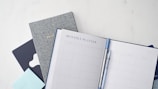 A neatly arranged stationery setup featuring a monthly planner open to a blank calendar layout, accompanied by a blue pen clipped onto it. Behind the planner, there are additional books or notebooks, including a gray one labeled 'Bullet' and another dark blue item. A light blue sticky note pad is also visible.