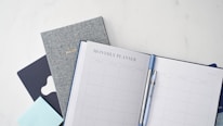 A neatly arranged stationery setup featuring a monthly planner open to a blank calendar layout, accompanied by a blue pen clipped onto it. Behind the planner, there are additional books or notebooks, including a gray one labeled 'Bullet' and another dark blue item. A light blue sticky note pad is also visible.