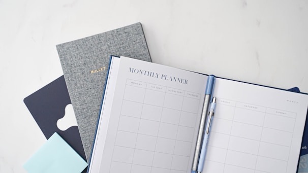 A neatly arranged stationery setup featuring a monthly planner open to a blank calendar layout, accompanied by a blue pen clipped onto it. Behind the planner, there are additional books or notebooks, including a gray one labeled 'Bullet' and another dark blue item. A light blue sticky note pad is also visible.