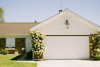 A charming single-story house with a white garage door and a sloped red-tiled roof. The facade includes a small garden with yellow flowers climbing on both sides of the garage. The sky is clear and blue, creating a serene atmosphere.