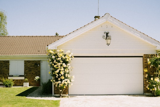 A charming single-story house with a white garage door and a sloped red-tiled roof. The facade includes a small garden with yellow flowers climbing on both sides of the garage. The sky is clear and blue, creating a serene atmosphere.