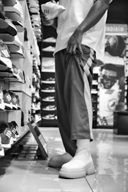 A person is standing in a shoe store, holding a sneaker while looking at a wall full of shoes. The scene is in black and white, with the person wearing casual pants and shoes. Various sneakers are displayed on shelves, arranged neatly in rows.
