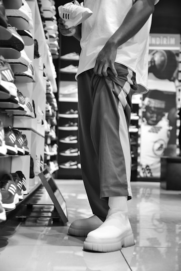 A person is standing in a shoe store, holding a sneaker while looking at a wall full of shoes. The scene is in black and white, with the person wearing casual pants and shoes. Various sneakers are displayed on shelves, arranged neatly in rows.