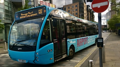 A blue airport express bus is parked on a city street near a modern glass building. The destination board above the windshield displays the route information. A red no entry sign is visible in the foreground, and the street is bordered by modern buildings and trees.
