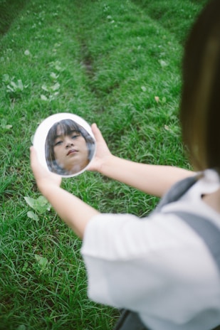 A child holds a circular mirror against a backdrop of lush green grass. The mirror reflects their face, showing a direct and introspective expression.