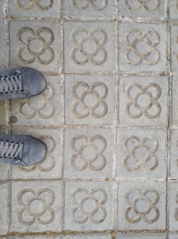 A patterned concrete pavement with floral-shaped motifs. Two sneakers are partially visible at the bottom left corner, suggesting someone standing on the tiles.