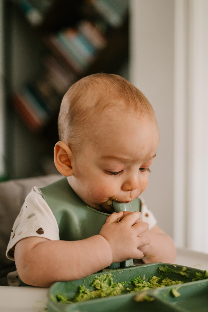 Baby sitting in a high chair eating solid food, making a slightly messy but happy face