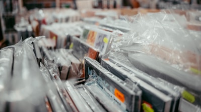A collection of framed pictures or artwork are wrapped in protective plastic. They are closely stacked, with some yellow and orange price tags visible. The setting appears to be a store or warehouse.