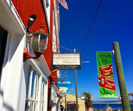 A street scene featuring red and white buildings with a vintage lantern and a sign for Steinbeck Jewelers. An American flag is visible on the building, and a green Cannery Row banner hangs from a pole. Additional signs for nearby establishments and a view of the ocean in the distance complete the vibrant setting.