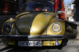 A vintage yellow car, likely a Volkswagen Beetle, is parked on a street. The car has a shiny surface with reflective lighting and a Turkish flag attached to the right side mirror. The background shows an orange vehicle and some trees, with buildings partially visible.