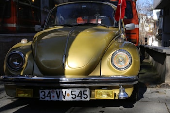A vintage yellow car, likely a Volkswagen Beetle, is parked on a street. The car has a shiny surface with reflective lighting and a Turkish flag attached to the right side mirror. The background shows an orange vehicle and some trees, with buildings partially visible.