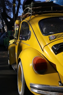 A classic yellow Volkswagen Beetle is parked on the street, with a roof rack carrying various items such as a green mat and others indistinguishable. The car is in good condition, with its shiny surface reflecting the daylight. In the background, part of another car is visible, possibly another Beetle, suggesting a gathering of vintage cars.