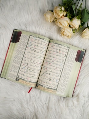 An open book with Arabic script is placed on a soft, white fur surface. Beside the book, there are several pale cream-colored roses with green leaves.