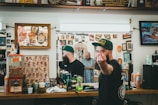 A barbershop setting with framed artwork and memorabilia on the walls. A bearded man wearing a green cap is giving a rude gesture towards the camera while looking reflected in the mirror. The countertop is cluttered with various grooming products, soda, and a gumball machine. Another person is visible in the mirror, seemingly focused on a task.