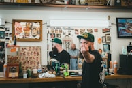 A barbershop setting with framed artwork and memorabilia on the walls. A bearded man wearing a green cap is giving a rude gesture towards the camera while looking reflected in the mirror. The countertop is cluttered with various grooming products, soda, and a gumball machine. Another person is visible in the mirror, seemingly focused on a task.