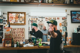 A barbershop setting with framed artwork and memorabilia on the walls. A bearded man wearing a green cap is giving a rude gesture towards the camera while looking reflected in the mirror. The countertop is cluttered with various grooming products, soda, and a gumball machine. Another person is visible in the mirror, seemingly focused on a task.