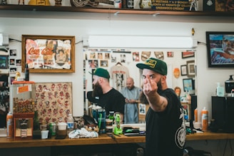 A barbershop setting with framed artwork and memorabilia on the walls. A bearded man wearing a green cap is giving a rude gesture towards the camera while looking reflected in the mirror. The countertop is cluttered with various grooming products, soda, and a gumball machine. Another person is visible in the mirror, seemingly focused on a task.