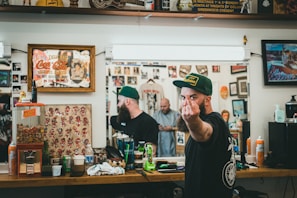 A barbershop setting with framed artwork and memorabilia on the walls. A bearded man wearing a green cap is giving a rude gesture towards the camera while looking reflected in the mirror. The countertop is cluttered with various grooming products, soda, and a gumball machine. Another person is visible in the mirror, seemingly focused on a task.