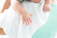 A close-up of a baby's hand resting on a white cloth. The infant's fingers are relaxed, and the skin appears soft and smooth. The background features a soft teal hue, indicating a calm and serene setting.