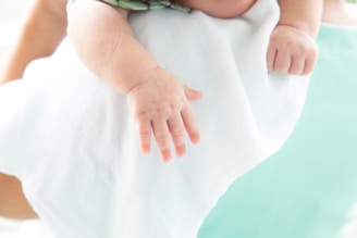 A close-up of a baby's hand resting on a white cloth. The infant's fingers are relaxed, and the skin appears soft and smooth. The background features a soft teal hue, indicating a calm and serene setting.
