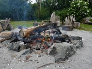 A campfire pit with charred logs and glowing embers surrounded by rocks. The area is sandy, with rustic wooden chairs in the background, set in a lush green garden with flowering bushes.