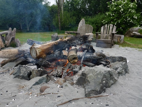 A campfire pit with charred logs and glowing embers surrounded by rocks. The area is sandy, with rustic wooden chairs in the background, set in a lush green garden with flowering bushes.