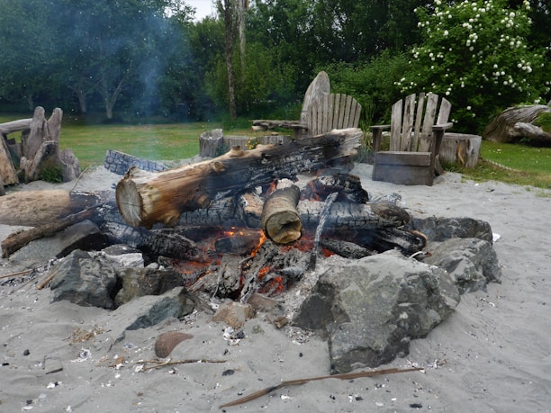A campfire pit with charred logs and glowing embers surrounded by rocks. The area is sandy, with rustic wooden chairs in the background, set in a lush green garden with flowering bushes.