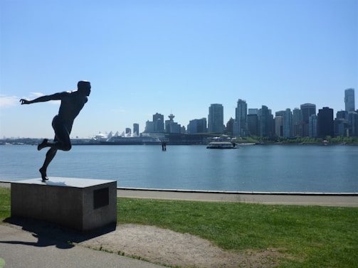 A bronze statue of a runner is positioned on a pedestal along a waterfront, with a modern city skyline visible across the water under a clear blue sky.