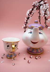 A whimsical teapot and teacup with smiling faces are positioned against a soft pink background. The teapot has a purple lid, while the teacup features a golden handle. Delicate pink cherry blossoms are arranged around and arching over the teapot, with a scattering of petals on the surface.