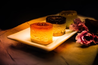 Three round, layered desserts are elegantly presented on a rectangular white plate. The desserts vary in color, with one featuring a caramel-colored top, another with a dark chocolate layer, and the third hinting a lighter topping. A set of pink flowers lies aesthetically arranged beside the plate, with warm lighting casting a cozy, inviting glow over the scene.