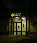 The entrance of a bank is illuminated at night. The bank name, 'Halyk Bank,' is prominently displayed with bright green signage. A digital display shows red numbers to the left of the entrance. The glass doors and windows reveal a well-lit interior environment, and an orange traffic cone is visible near the doors.