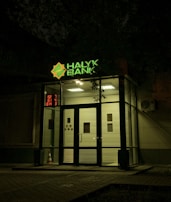 The entrance of a bank is illuminated at night. The bank name, 'Halyk Bank,' is prominently displayed with bright green signage. A digital display shows red numbers to the left of the entrance. The glass doors and windows reveal a well-lit interior environment, and an orange traffic cone is visible near the doors.