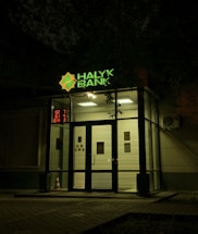 The entrance of a bank is illuminated at night. The bank name, 'Halyk Bank,' is prominently displayed with bright green signage. A digital display shows red numbers to the left of the entrance. The glass doors and windows reveal a well-lit interior environment, and an orange traffic cone is visible near the doors.
