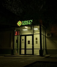 The entrance of a bank is illuminated at night. The bank name, 'Halyk Bank,' is prominently displayed with bright green signage. A digital display shows red numbers to the left of the entrance. The glass doors and windows reveal a well-lit interior environment, and an orange traffic cone is visible near the doors.