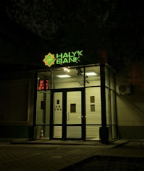 The entrance of a bank is illuminated at night. The bank name, 'Halyk Bank,' is prominently displayed with bright green signage. A digital display shows red numbers to the left of the entrance. The glass doors and windows reveal a well-lit interior environment, and an orange traffic cone is visible near the doors.