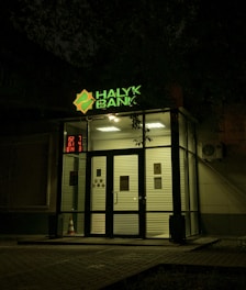 The entrance of a bank is illuminated at night. The bank name, 'Halyk Bank,' is prominently displayed with bright green signage. A digital display shows red numbers to the left of the entrance. The glass doors and windows reveal a well-lit interior environment, and an orange traffic cone is visible near the doors.