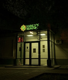 The entrance of a bank is illuminated at night. The bank name, 'Halyk Bank,' is prominently displayed with bright green signage. A digital display shows red numbers to the left of the entrance. The glass doors and windows reveal a well-lit interior environment, and an orange traffic cone is visible near the doors.
