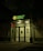 The entrance of a bank is illuminated at night. The bank name, 'Halyk Bank,' is prominently displayed with bright green signage. A digital display shows red numbers to the left of the entrance. The glass doors and windows reveal a well-lit interior environment, and an orange traffic cone is visible near the doors.