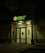 The entrance of a bank is illuminated at night. The bank name, 'Halyk Bank,' is prominently displayed with bright green signage. A digital display shows red numbers to the left of the entrance. The glass doors and windows reveal a well-lit interior environment, and an orange traffic cone is visible near the doors.